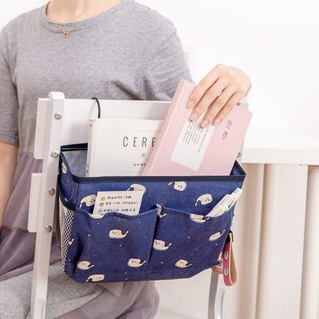 Blue bedside storage bag with multiple pockets holding books, stationery, and small items, hanging beside a chair, by Shopway.